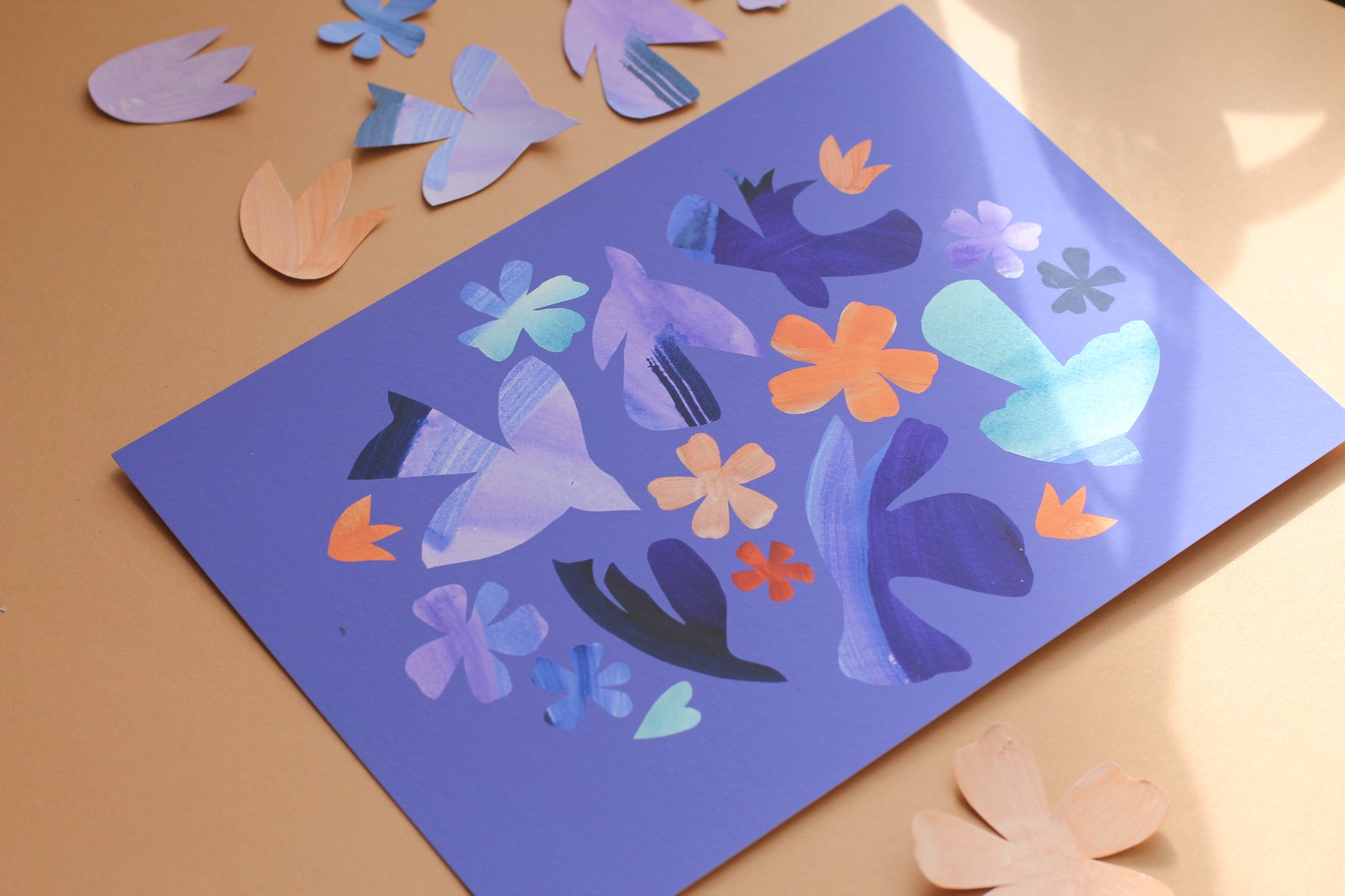 A bright colourful wall art print of simple bird and flower forms against a periwinkle blue background. The print is photographed against an earth toned background surrounded by paper cut outs of birds and flowers.