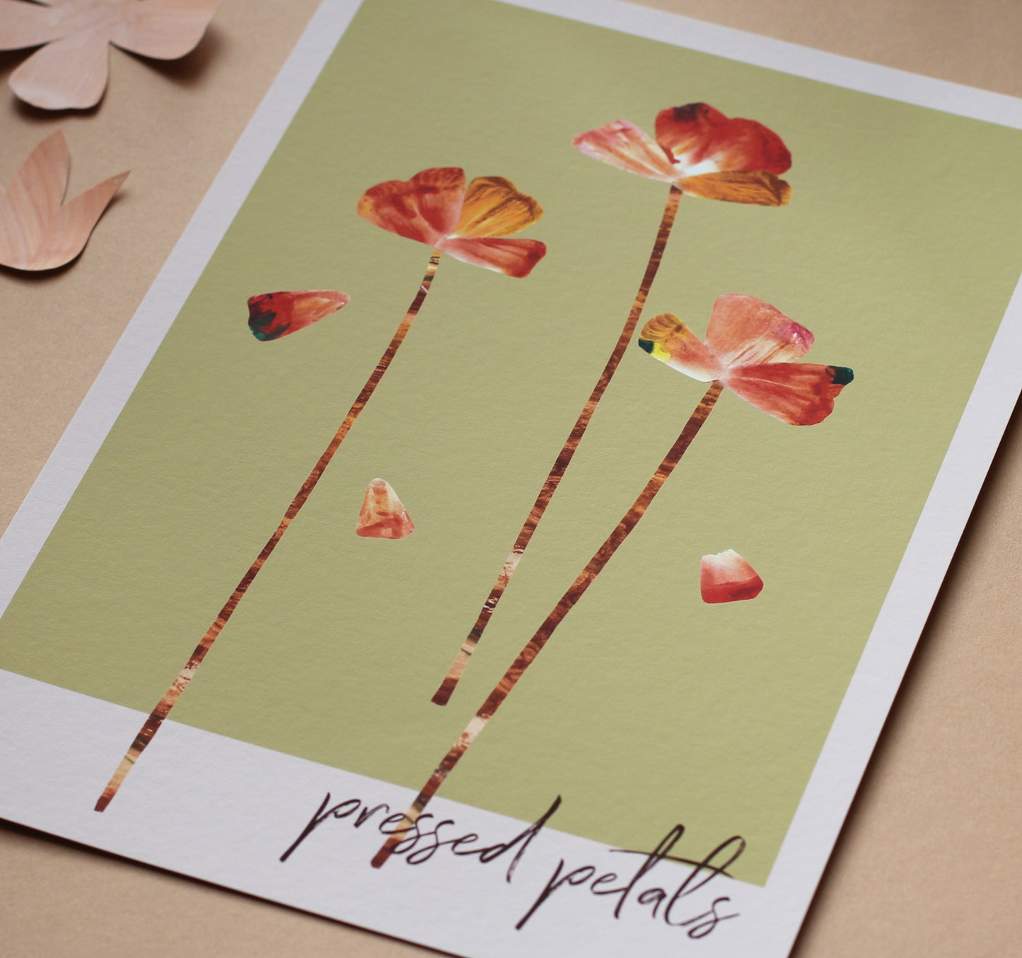 An art print of flower heads and petals in shades of orange, pink and rust against a soft green background. The words 'pressed petals' are written in script in the bottom right corner. The print is photographed unframed against a peachy background.