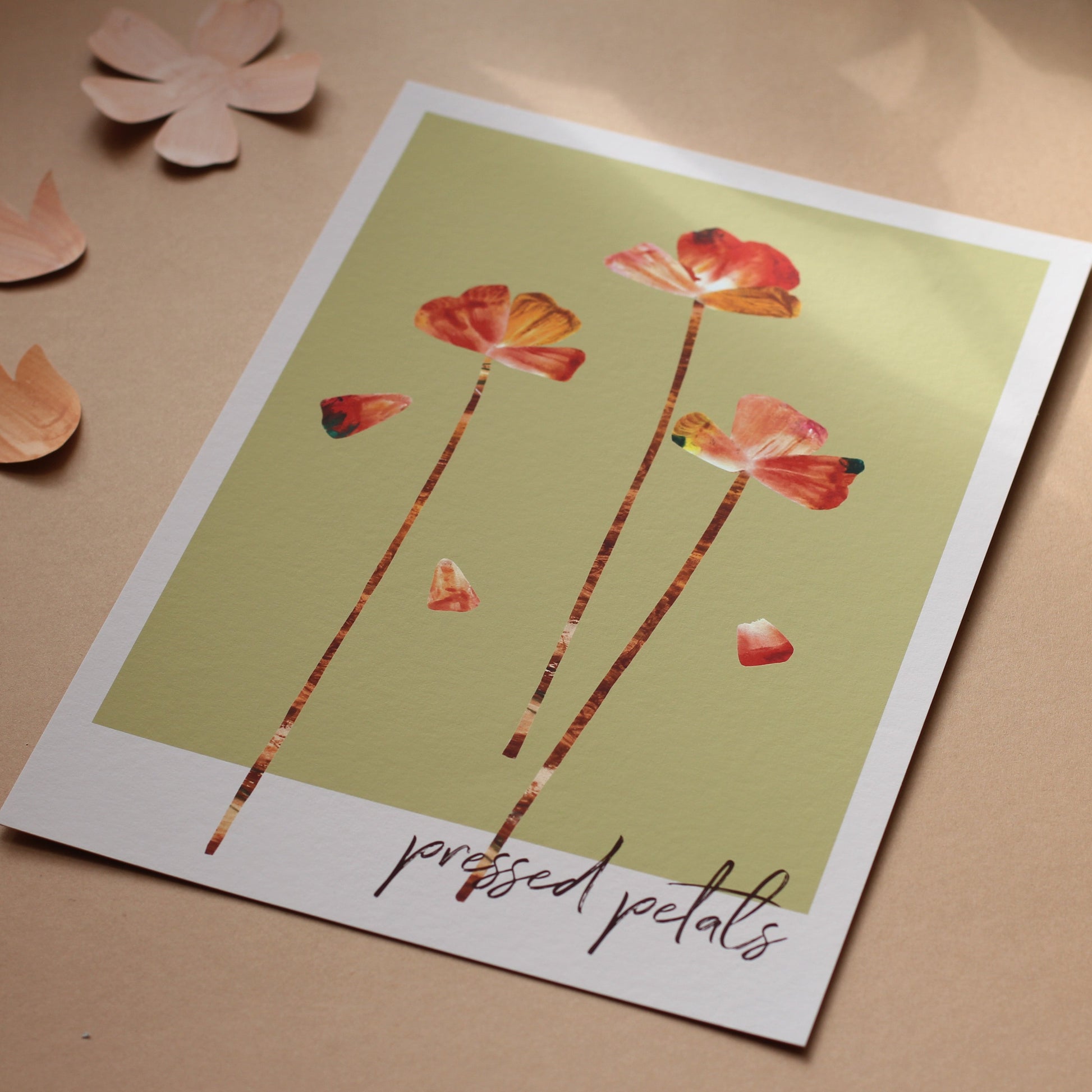 An art print of flower heads and petals in shades of orange, pink and rust against a soft green background. The words 'pressed petals' are written in script in the bottom right corner. The print is photographed unframed against a peachy background.