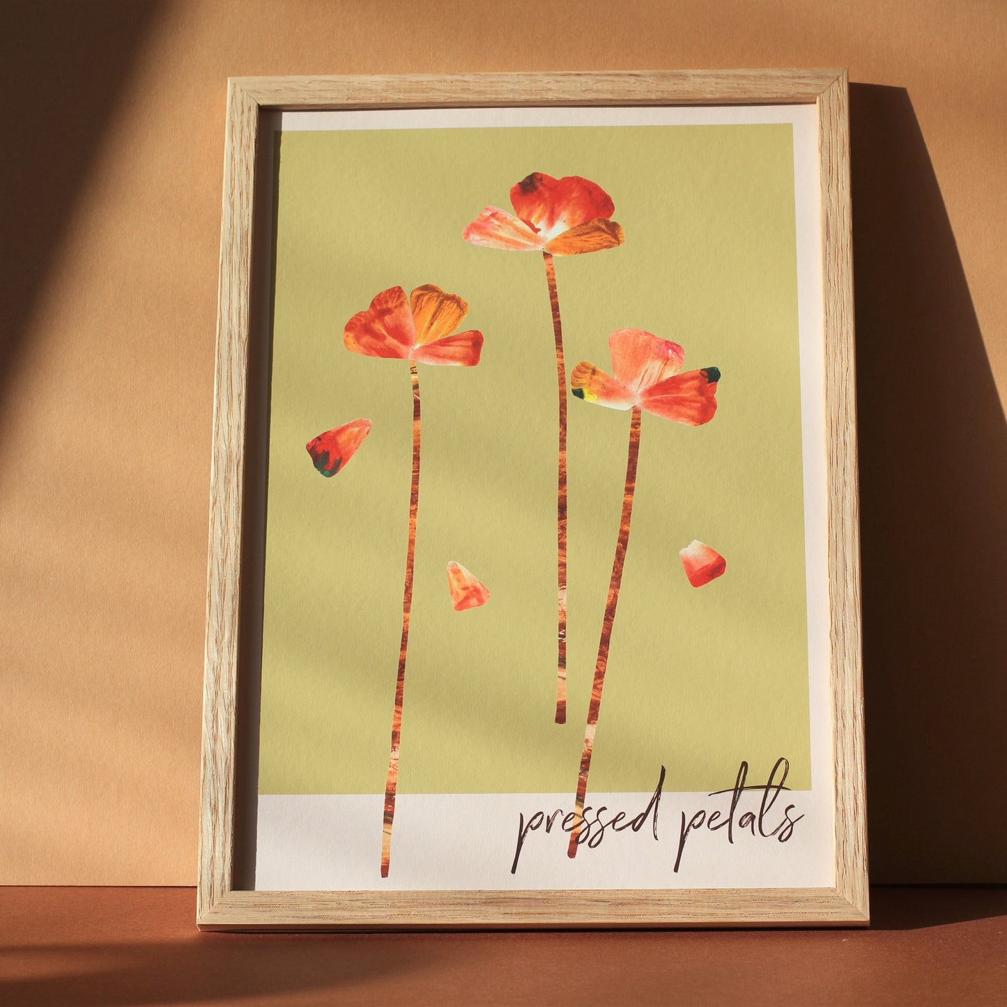 An art print of flower heads and petals in shades of orange, pink and rust against a soft green background. The words 'pressed petals' are written in script in the bottom right corner. The print is photographed framed against an earthy toned background.