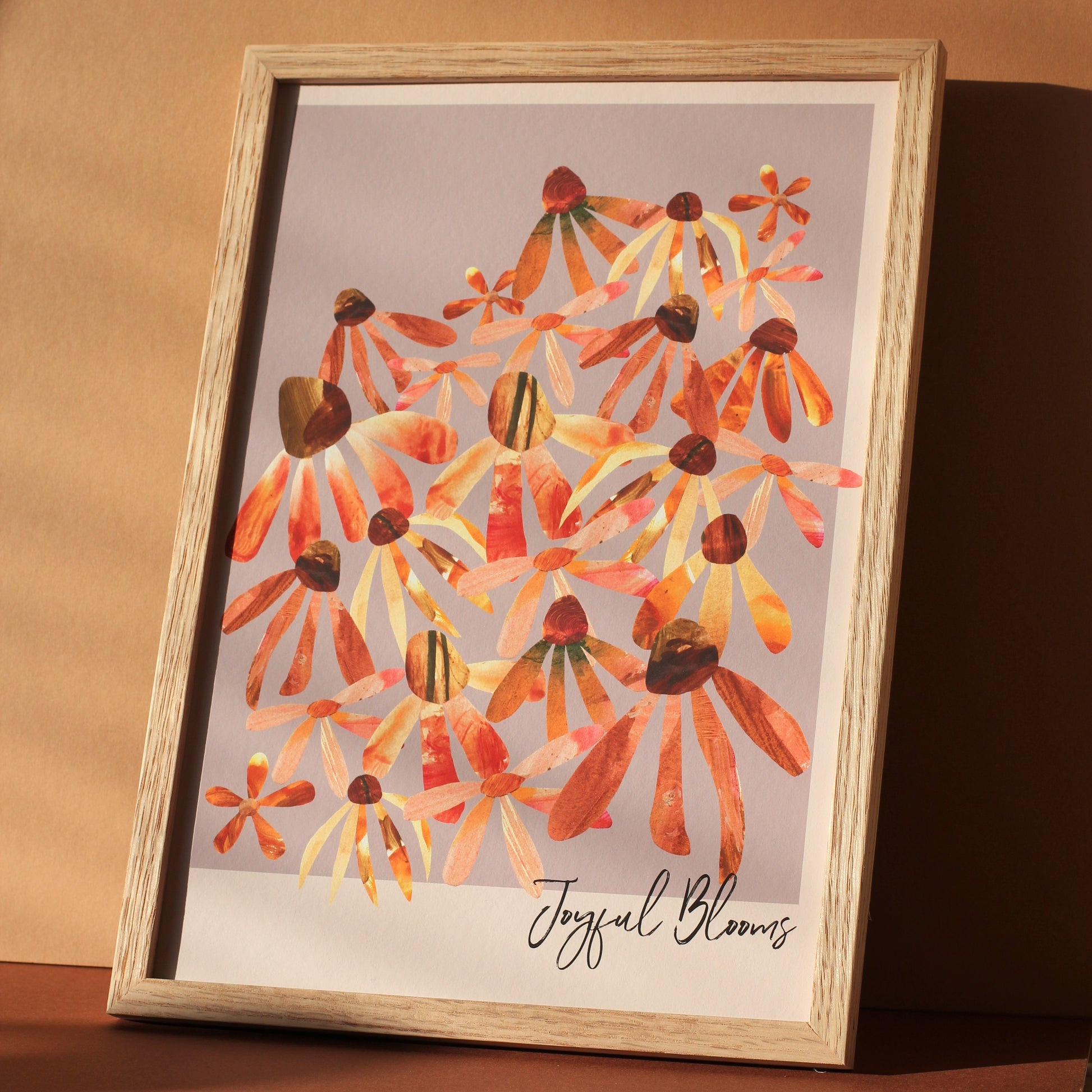 A floral art print with stylised daisy and echinacea flower heads in pinks, oranges and rusts against a soft lilac background. The words 'Joyful Blooms' are written in brush script in the bottom left hand corner of the print. The art print is photographed framed against a sunny wall.