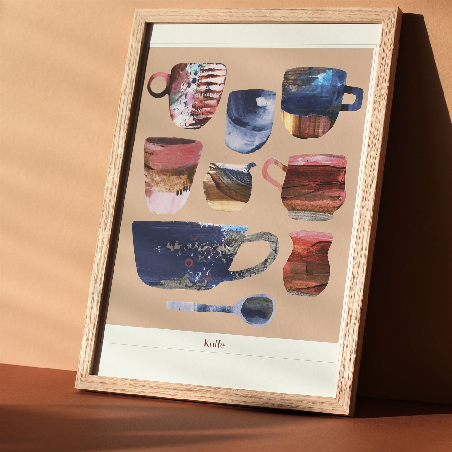 An art print for coffee lovers. The print shows a group of retro coffee cups, jugs and spoons in pinks, browns and china blues against a sand coloured background. The print is photographed framed in a sunny room.