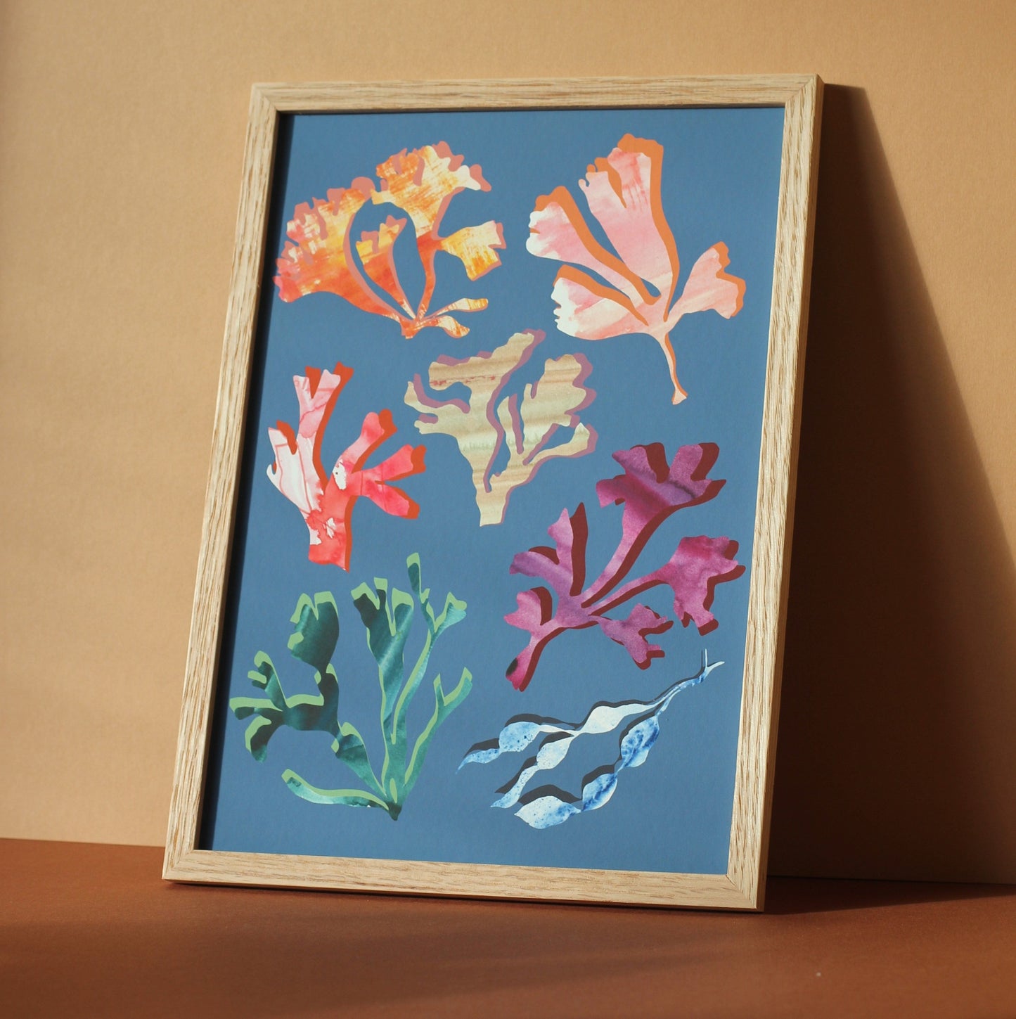 An art print of colourful seaweeds and corals against a mid blue background. The print is photographed in a wooden frame in a sunny room.