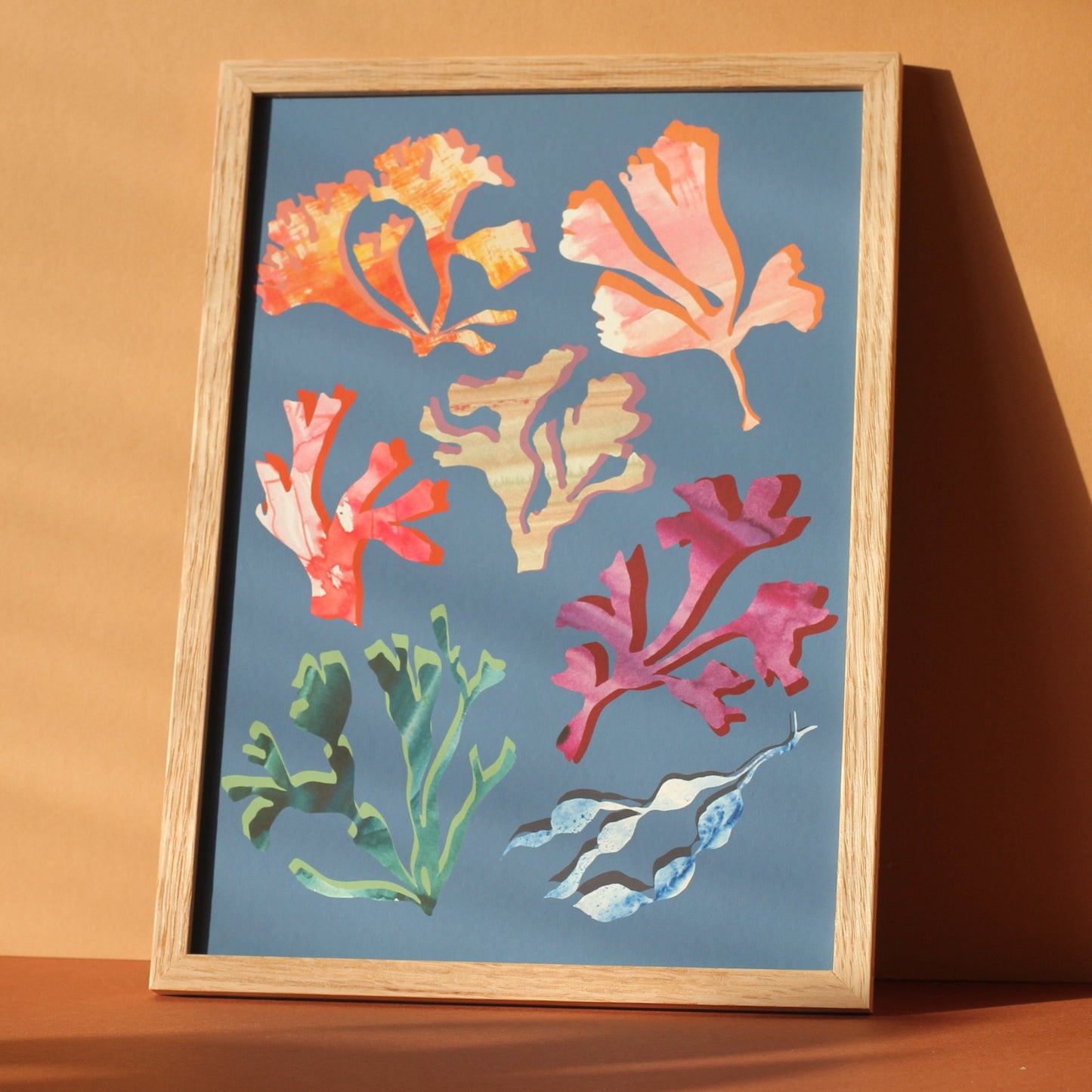 An art print of colourful seaweeds and corals against a mid blue background. The print is photographed in a wooden frame in a sunny room.