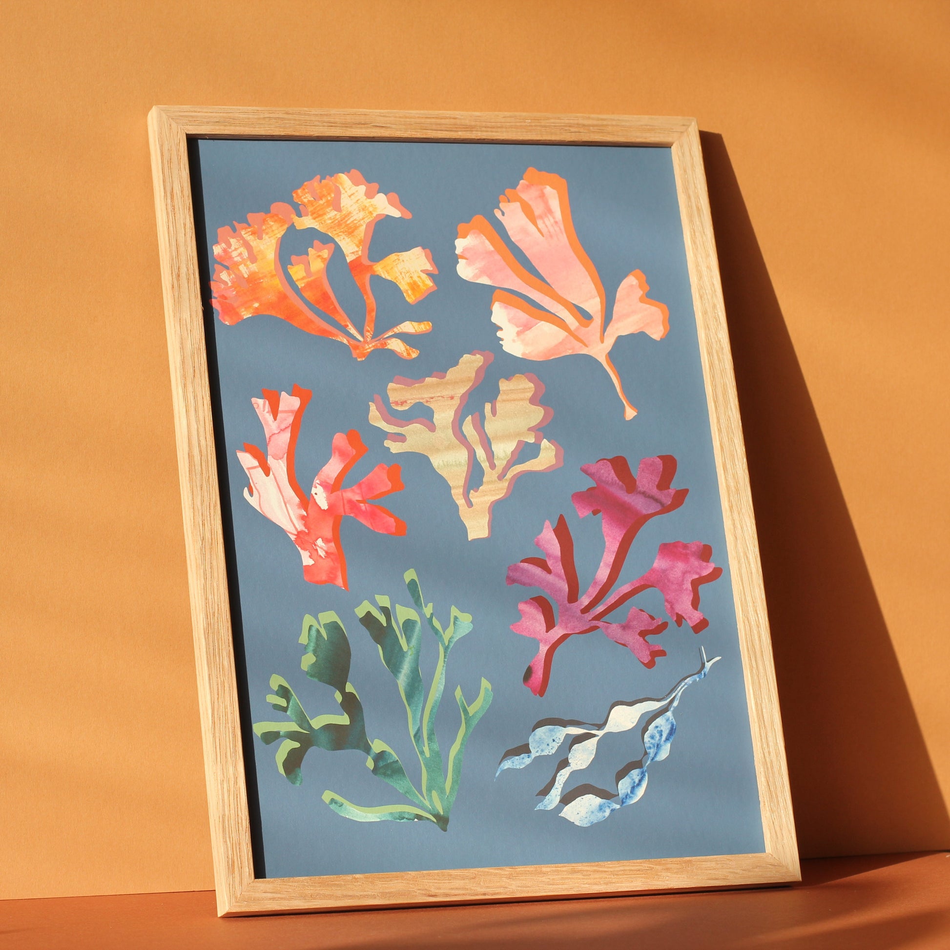 An art print of colourful seaweeds and corals against a mid blue background. The print is photographed in a wooden frame in a sunny room.