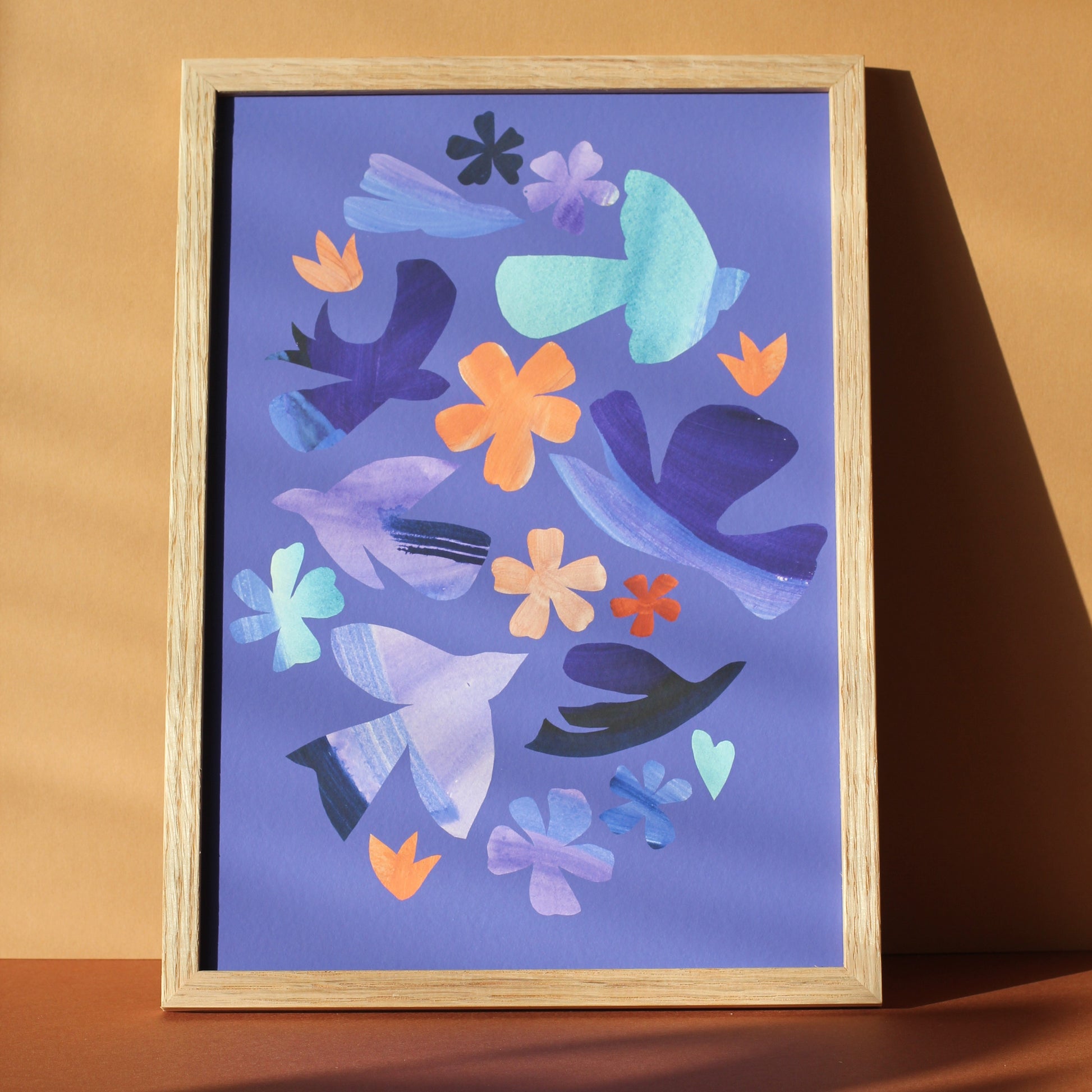 A bright colourful wall art print of simple bird and flower forms against a periwinkle blue background. The print is framed and photographed against an earth toned background.