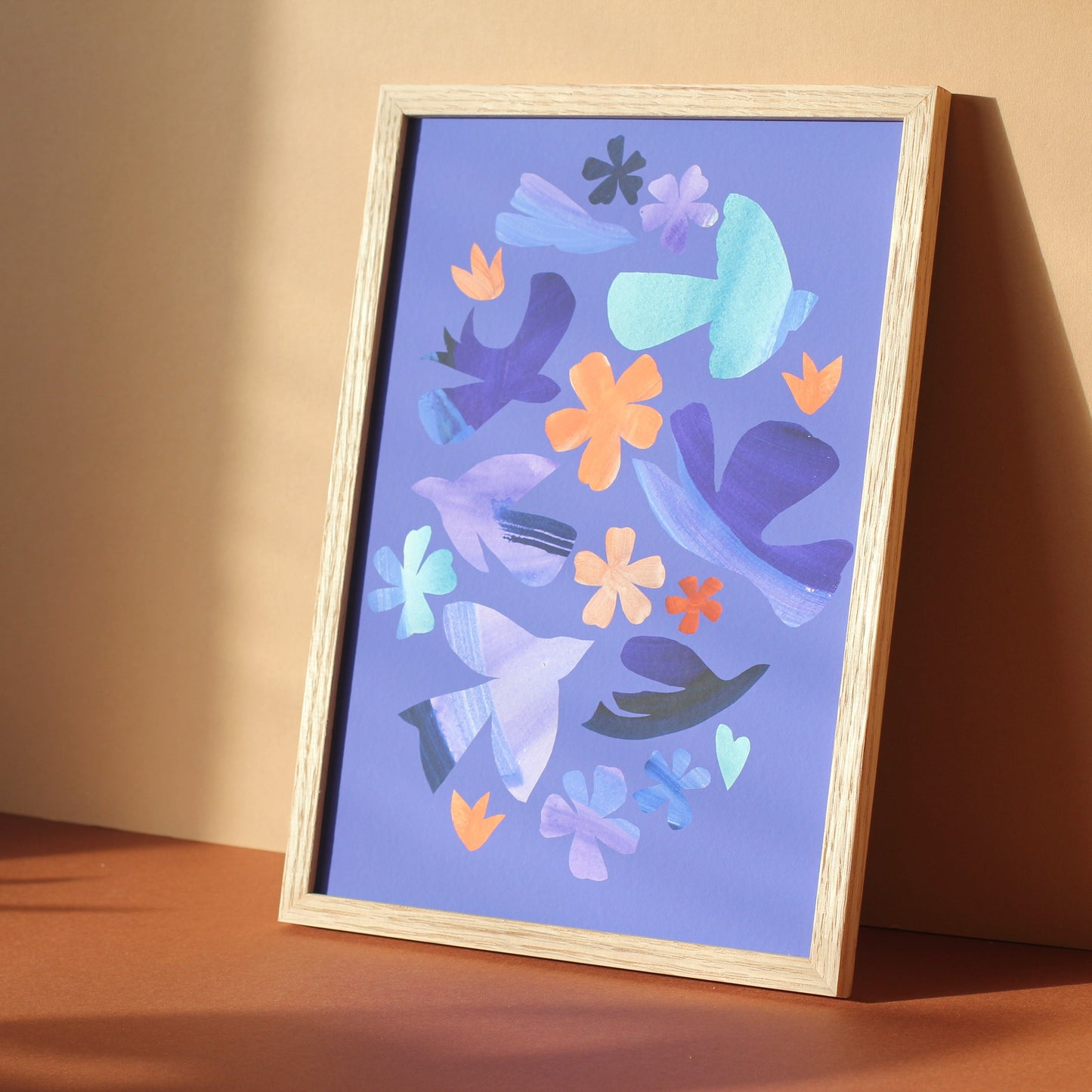 A bright colourful wall art print of simple bird and flower forms against a periwinkle blue background. The print is framed and photographed against an earth toned background.