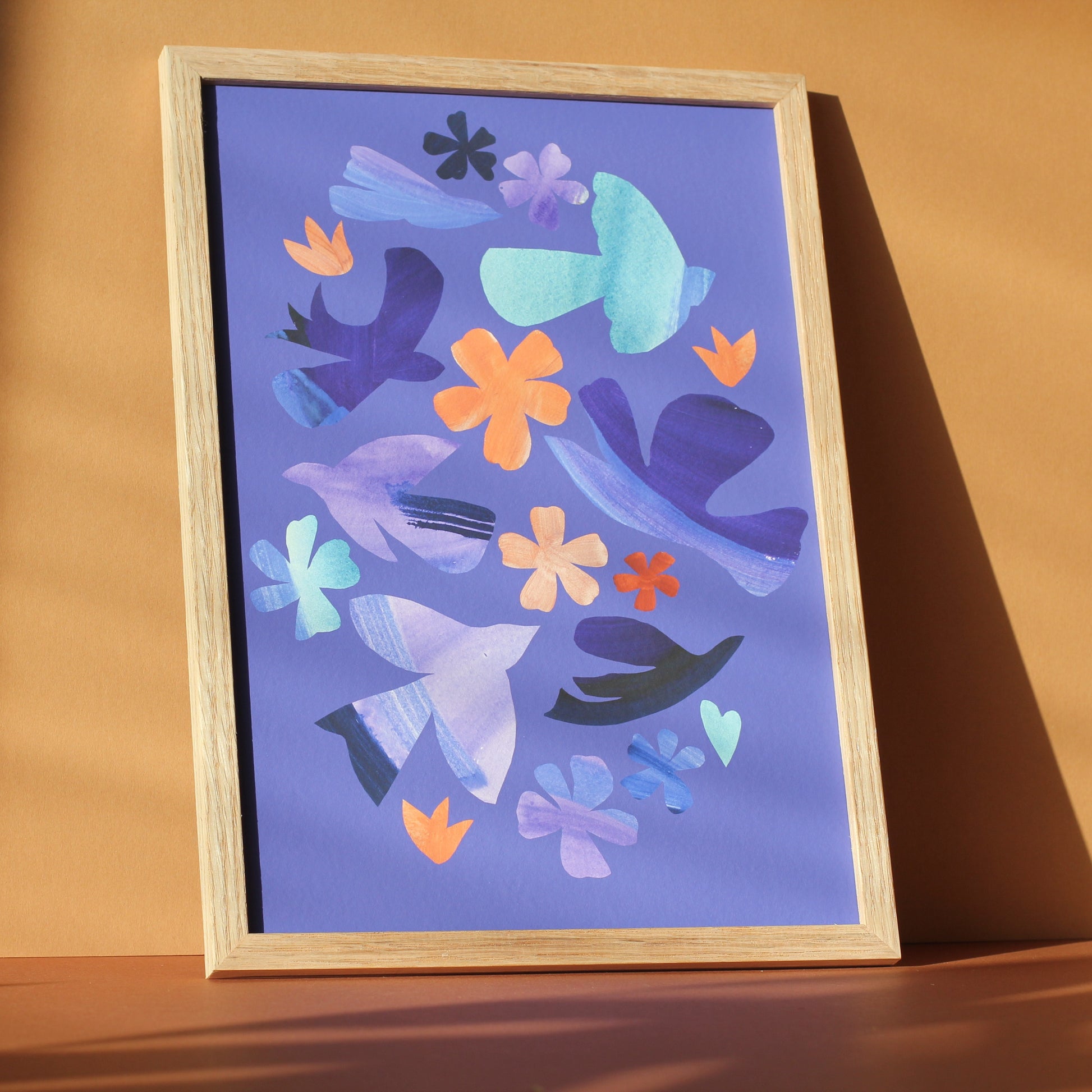 A bright colourful wall art print of simple bird and flower forms against a periwinkle blue background. The print is framed and photographed against an earth toned background.