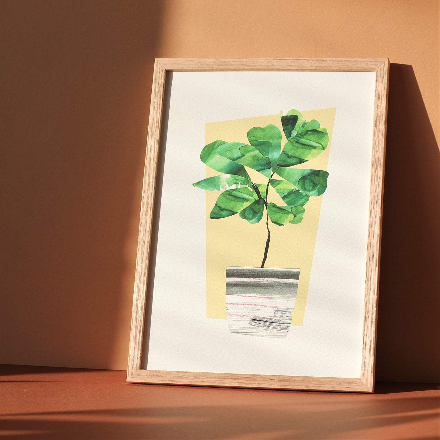 A houseplant art print of a fiddle leaf fig plant in a midcentury style planter. The original artwork was created using collaged watercolour papers. The print is photographed in a wooden frame against a sunny wall.