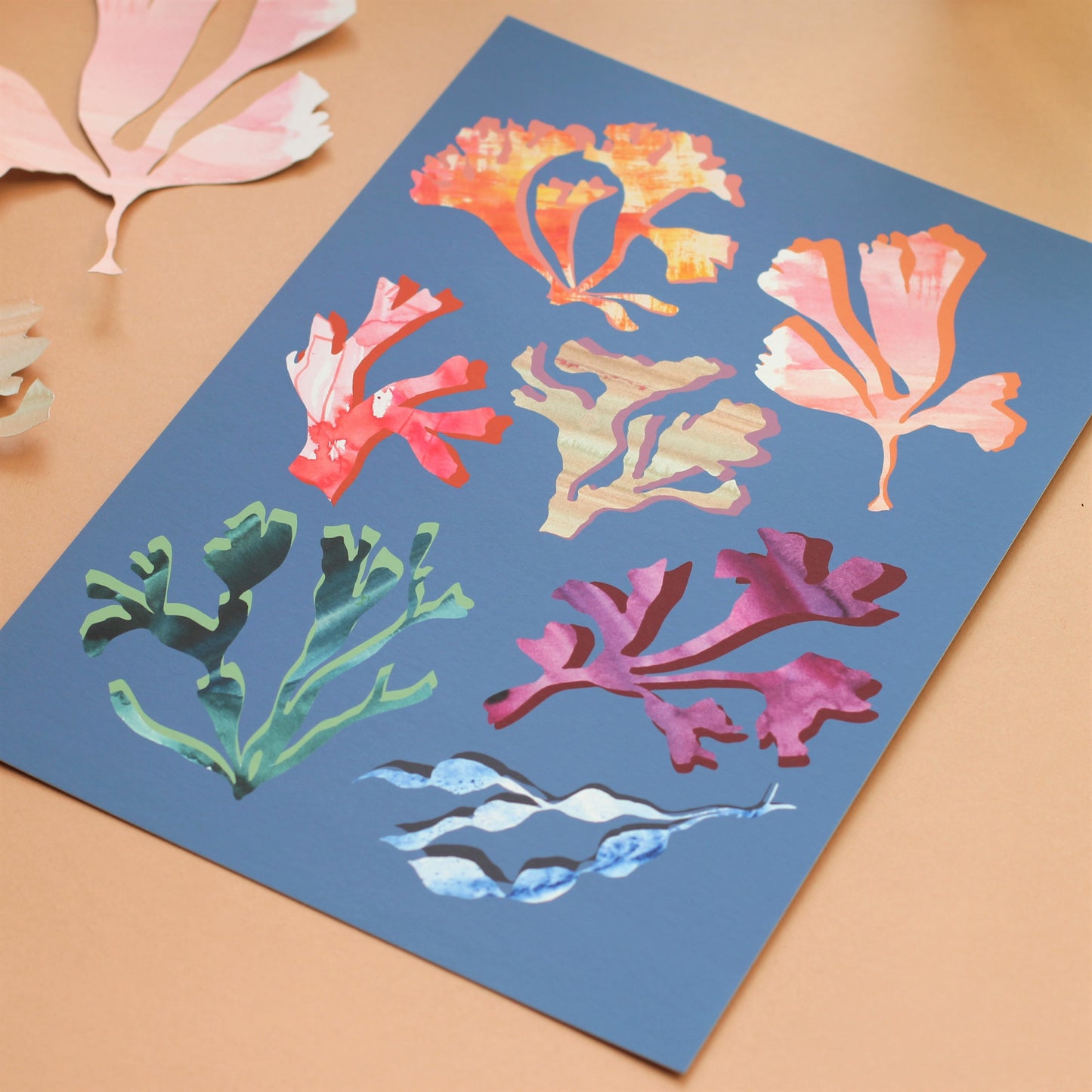 A colourful print of corals and seaweeds in a midcentury style. The corals and seaweeds are muted rainbow colours and the background is a mid blue.